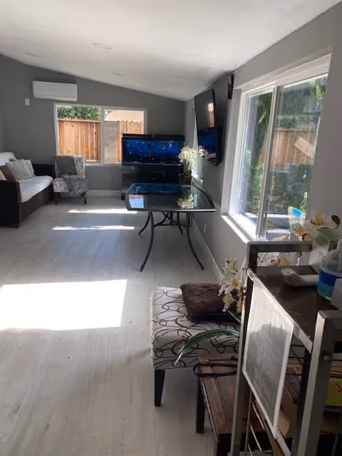 A bright living room with light-colored wooden flooring, a large window on the right letting in natural light, a glass table with a flower vase, a wall-mounted TV, a sofa and an armchair near a window, and a small shelving unit with various items.