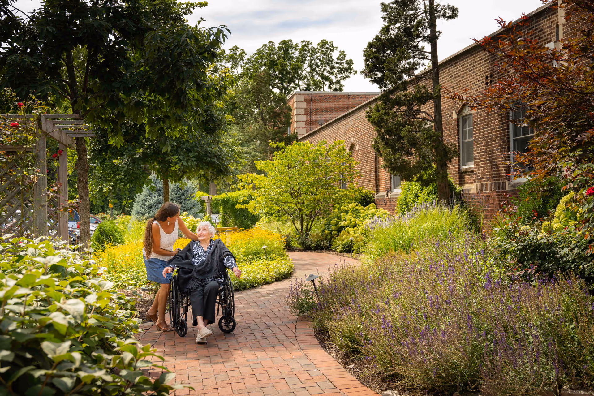A caregiver pushes an elderly woman in a wheelchair along a brick garden path beside a brick building surrounded by lush plants.