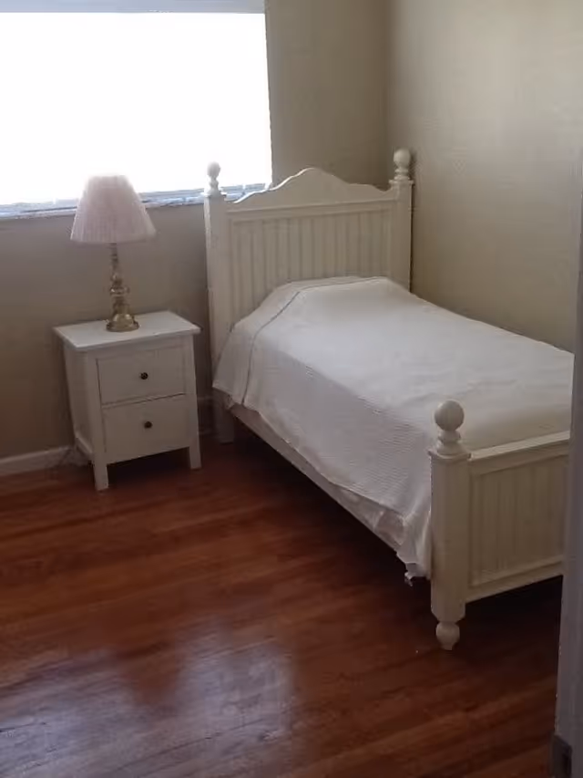 A small bedroom with a single white wooden bed covered with a white bedspread. Next to the bed is a white nightstand with two drawers and a lamp with a white lampshade. The room has wooden flooring and a window with blinds partially open, allowing natural light to enter.
