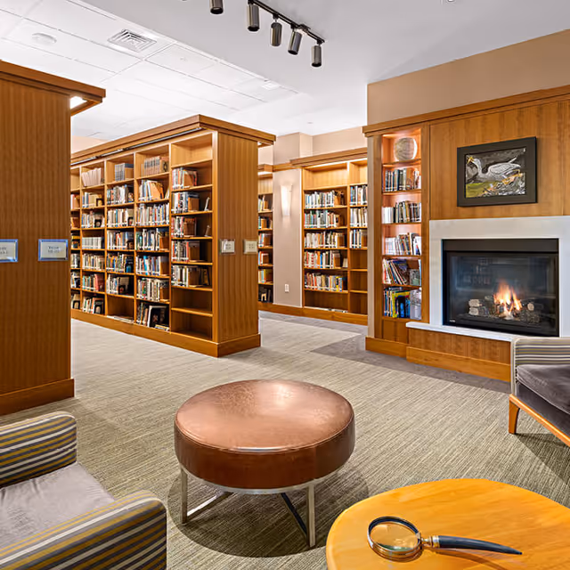 Cozy library reading area with wooden bookshelves, a lit fireplace, seating, and a round ottoman.