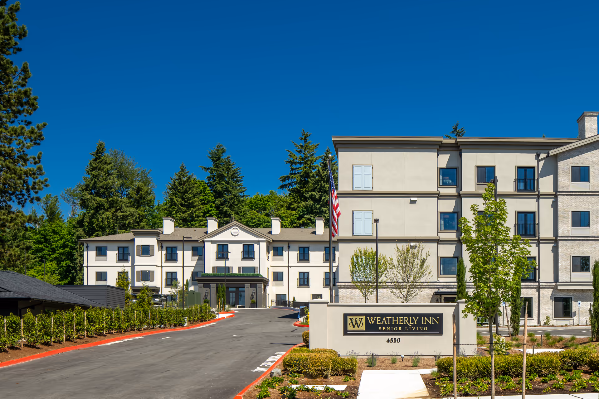 Exterior view of Weatherly Inn Renton senior living facility with a clear blue sky, a driveway leading to the entrance, landscaped greenery, and an American flag near the building sign.
