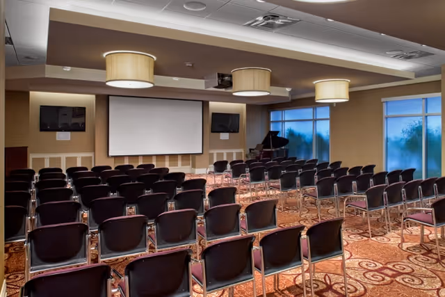 Carpeted meeting/auditorium room with rows of chairs facing a projection screen, wall monitors, pendant lights, and windows.