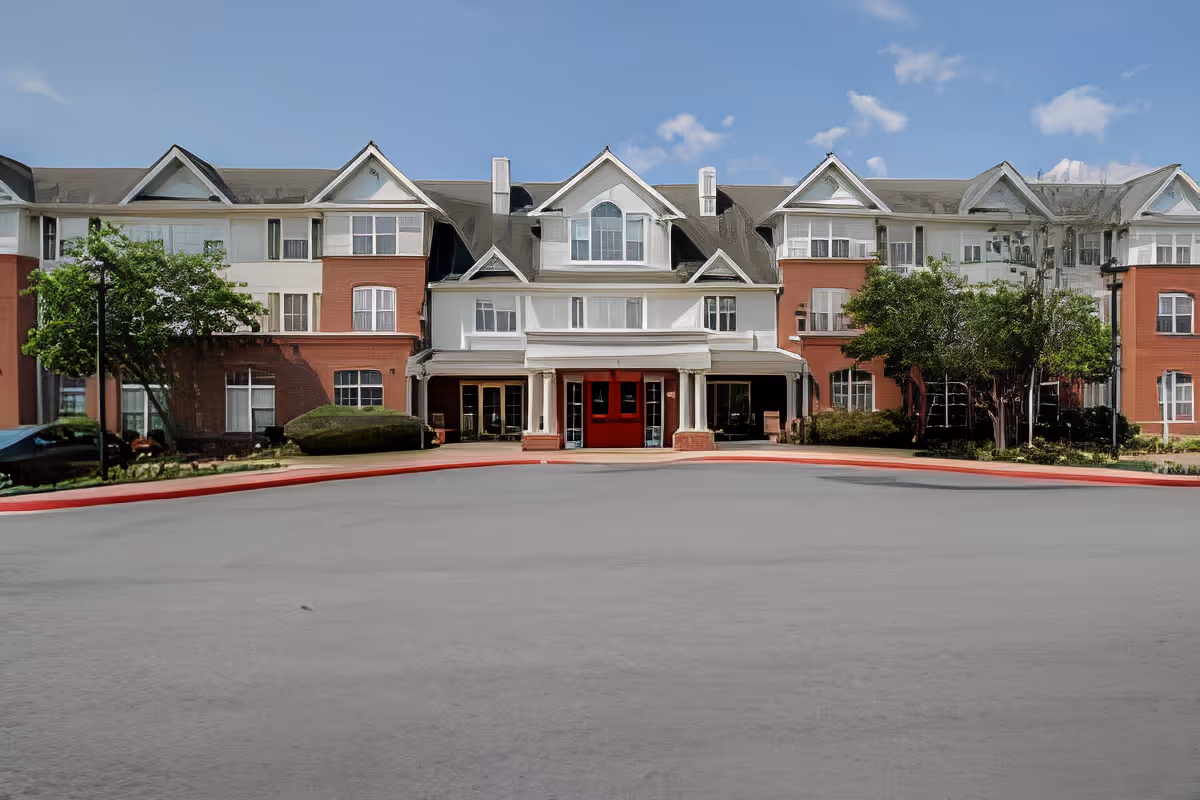 Front exterior view of Charter Senior Living of Woodholme Crossing, a large multi-story building with white and red brick facade, multiple windows, and a covered entrance with red double doors. The building is surrounded by trimmed bushes and trees under a blue sky with some clouds.