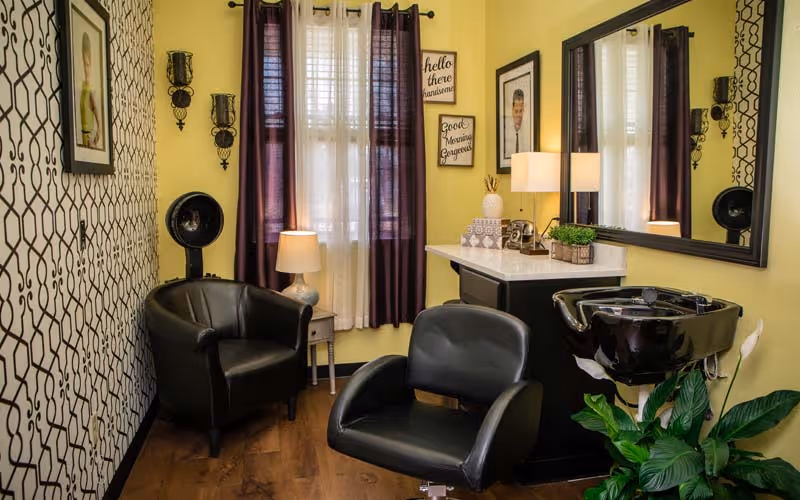 A cozy hair salon corner with two black salon chairs, a black hair washing sink, a small side table with a lamp, a large mirror on the wall, decorative wall art, and a window with dark purple and white curtains. The walls are painted yellow and one wall has a patterned wallpaper. There is also a green plant near the sink.