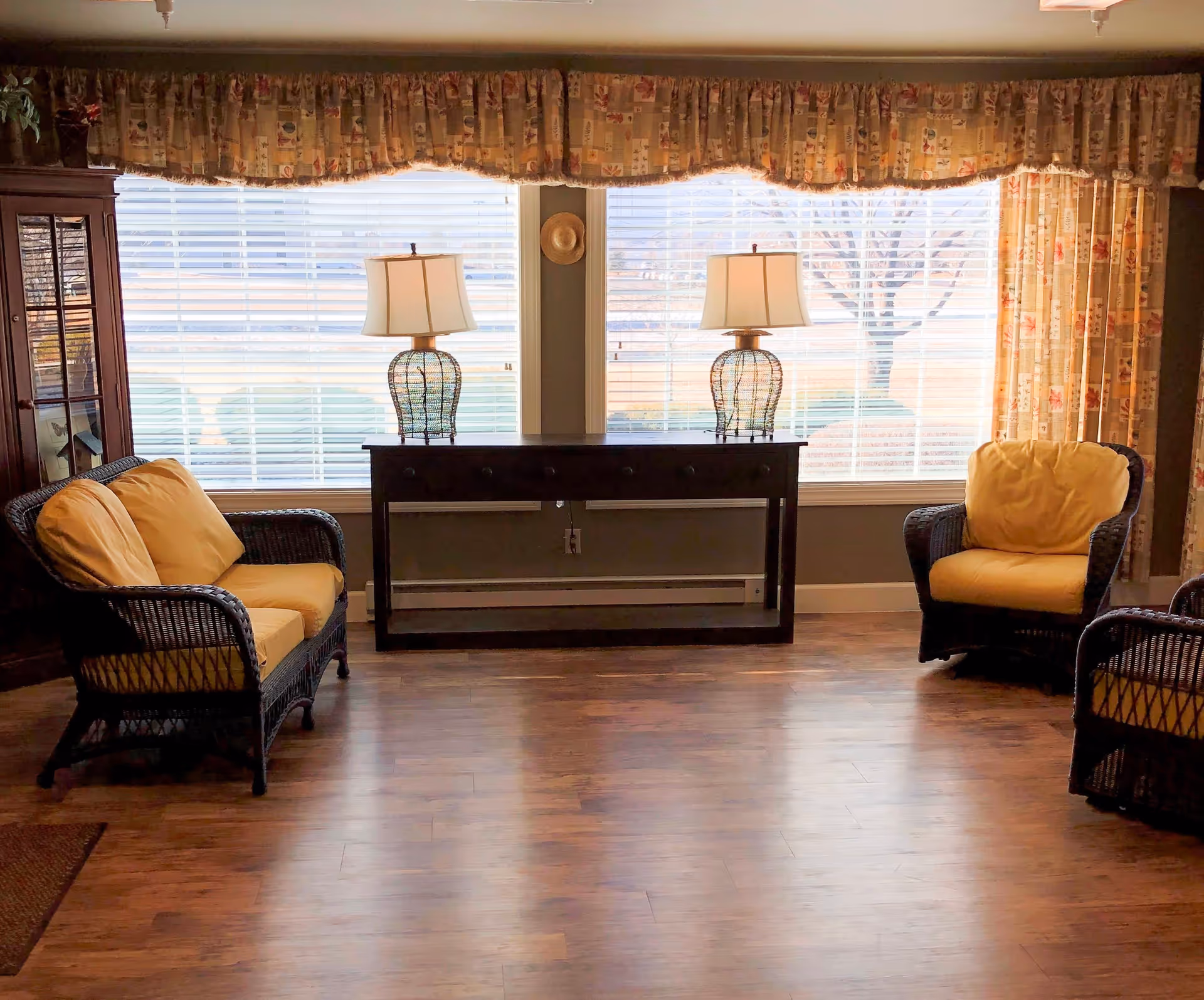 A cozy living room area with two wicker armchairs and a wicker loveseat, all with yellow cushions. A dark wooden console table with two decorative lamps is placed against a large window with white blinds and floral curtains. The floor is wooden, and there is a glass-front cabinet on the left side.