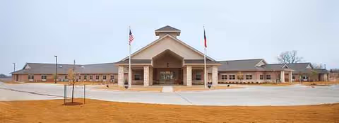 Front exterior of a single-story senior living facility with a covered entrance and flags.
