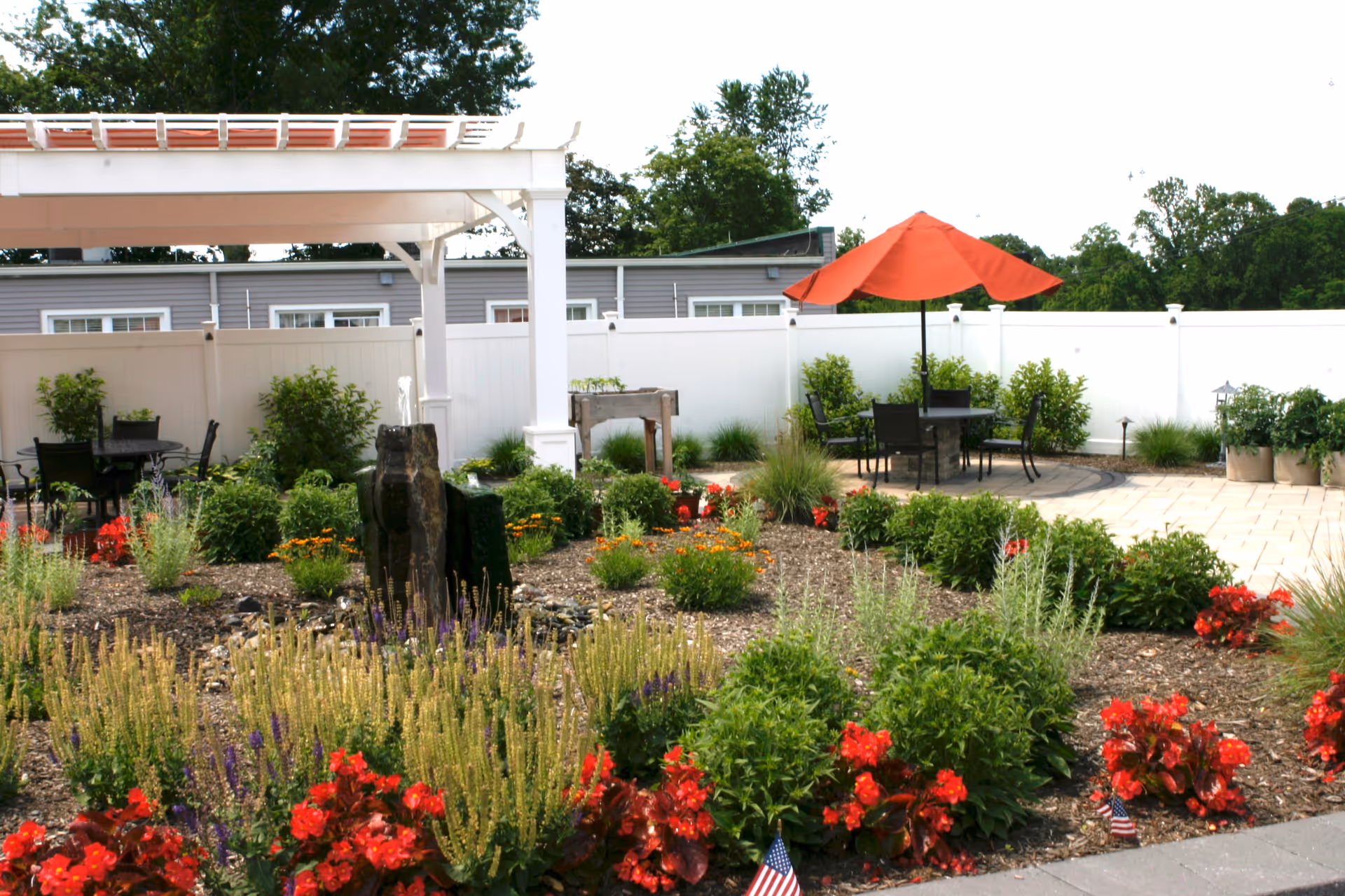 Outdoor courtyard with flowerbeds, a white pergola, patio tables and chairs under an orange umbrella.
