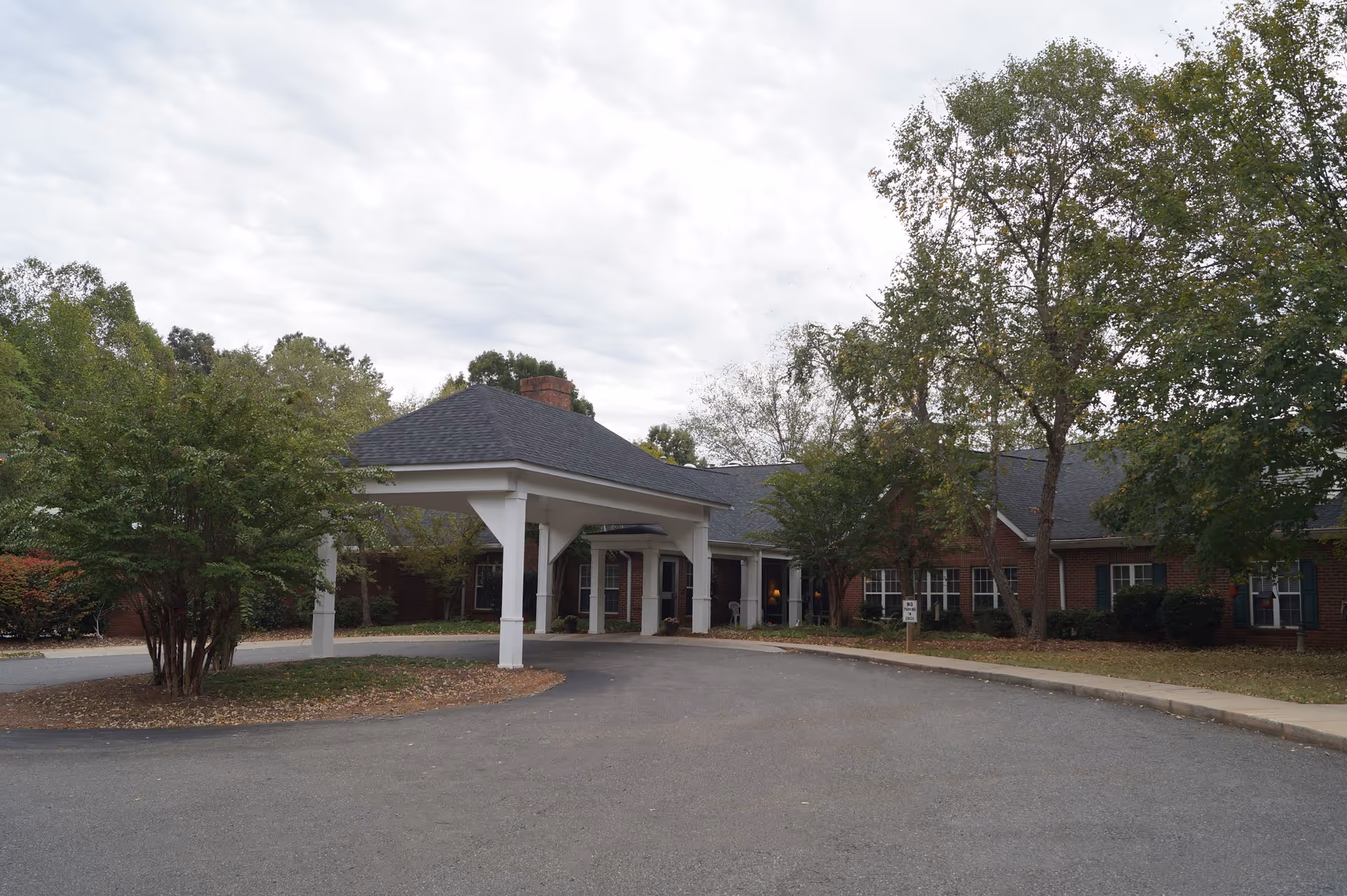 Front entrance of a single-story brick senior living building with a covered porte-cochere and surrounding trees.