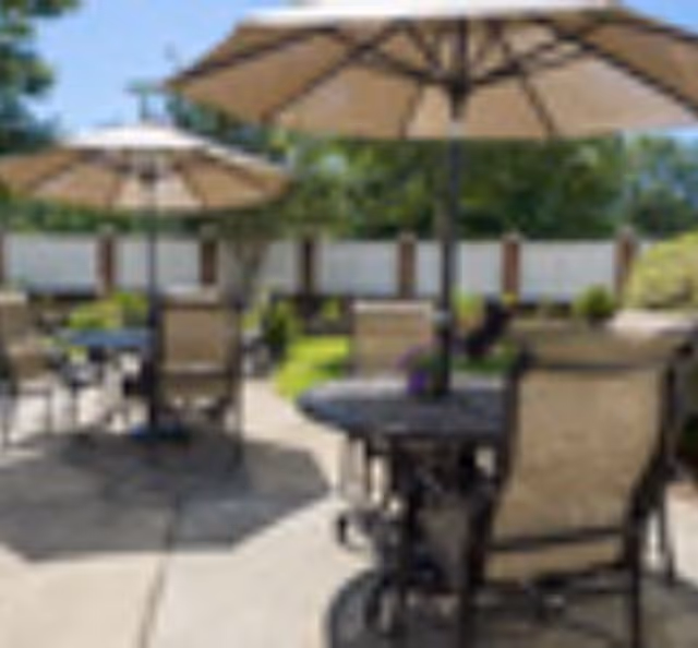 Outdoor patio with round tables, beige umbrellas, and cushioned chairs on a paved surface.