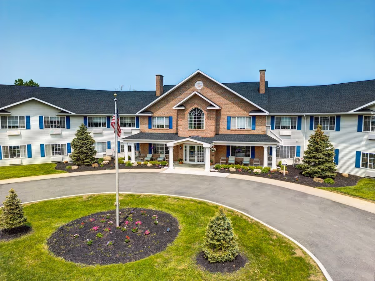 Front exterior view of Evergreen Place, a two-story senior living facility with white siding and blue shutters. The building has a central brick section with a large arched window above the entrance. A circular driveway surrounds a landscaped area with a flagpole and small trees.