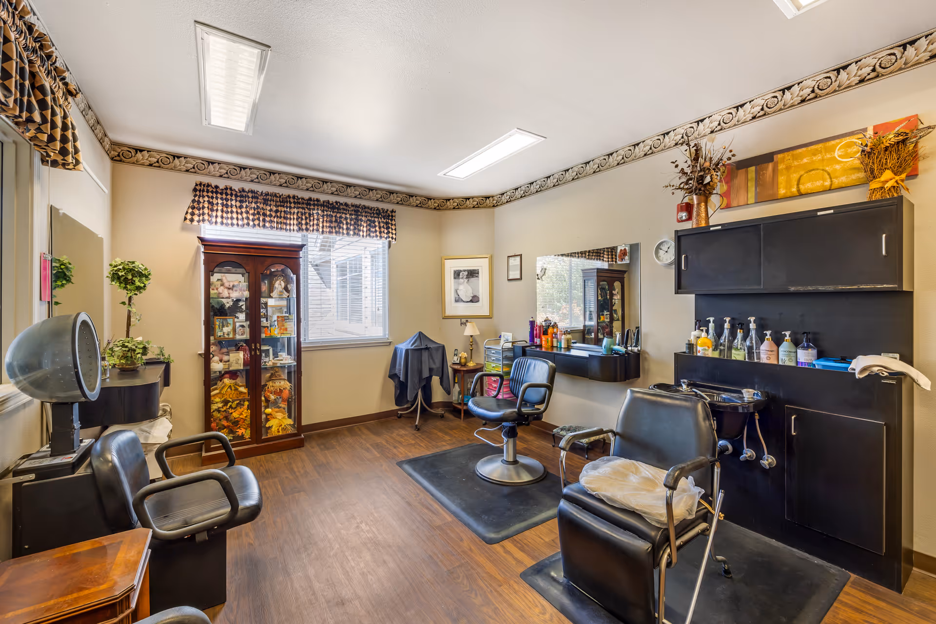 Interior of a hair salon room in a senior living facility with two black salon chairs, a hair washing station, a large mirror, and shelves with hair care products. The room has wood flooring, a window with patterned valance curtains, decorative trim near the ceiling, and a glass display cabinet with various decorative items.