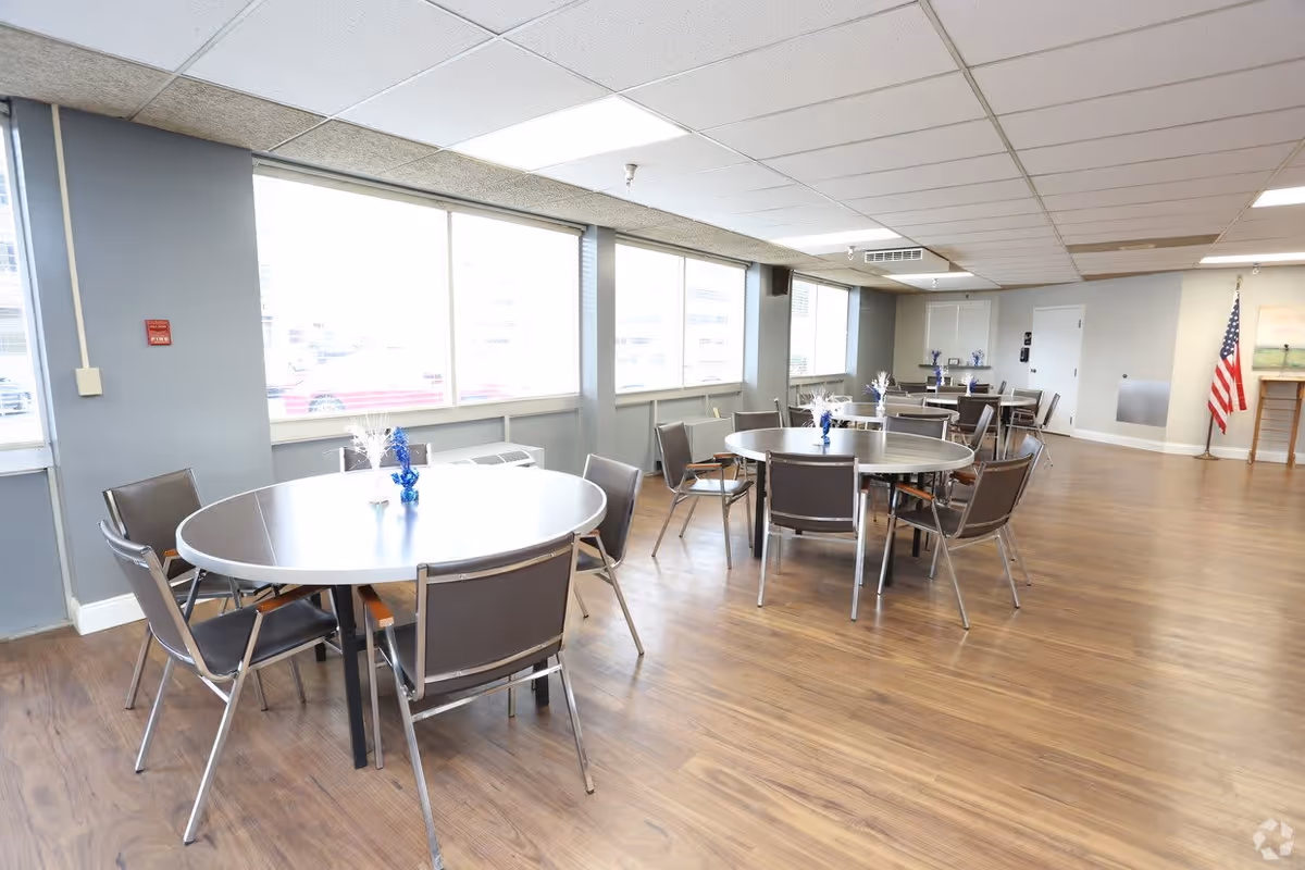 A spacious dining room with multiple round tables and chairs arranged neatly. The room has large windows allowing natural light to fill the space, wood flooring, and a ceiling with recessed lighting. An American flag is visible in the corner near a door.