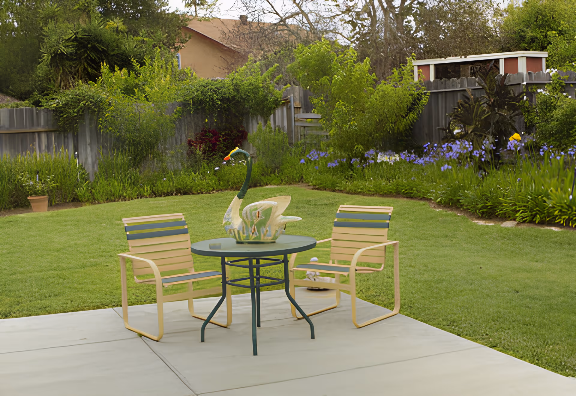 A peaceful outdoor garden area with a small round table and two chairs on a concrete patio. The table has a decorative swan sculpture on it. The garden is lush with green grass, various plants, flowers, and trees, enclosed by a wooden fence.