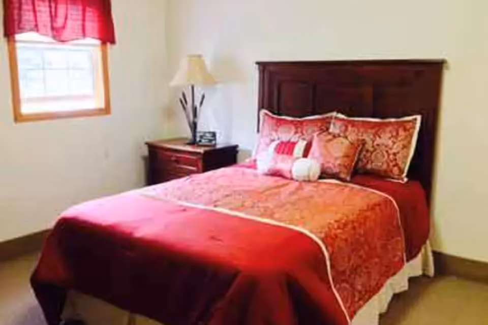 A bedroom with a wooden headboard bed dressed in red and patterned bedding, multiple decorative pillows, a wooden nightstand with a lamp, and a window with a red valance.