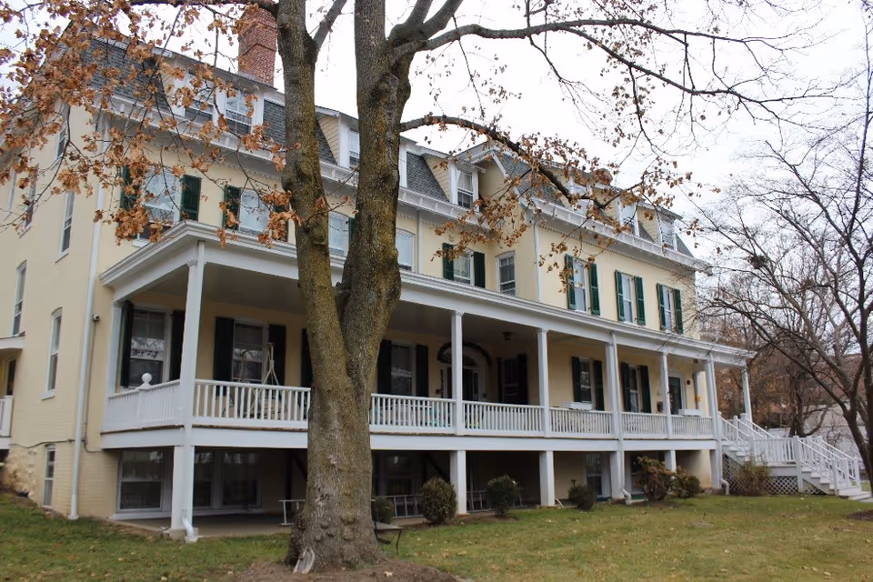 Exterior view of a large, multi-story beige building with green shutters and a white wrap-around porch. There are leafless trees in the foreground and a grassy lawn surrounding the building.