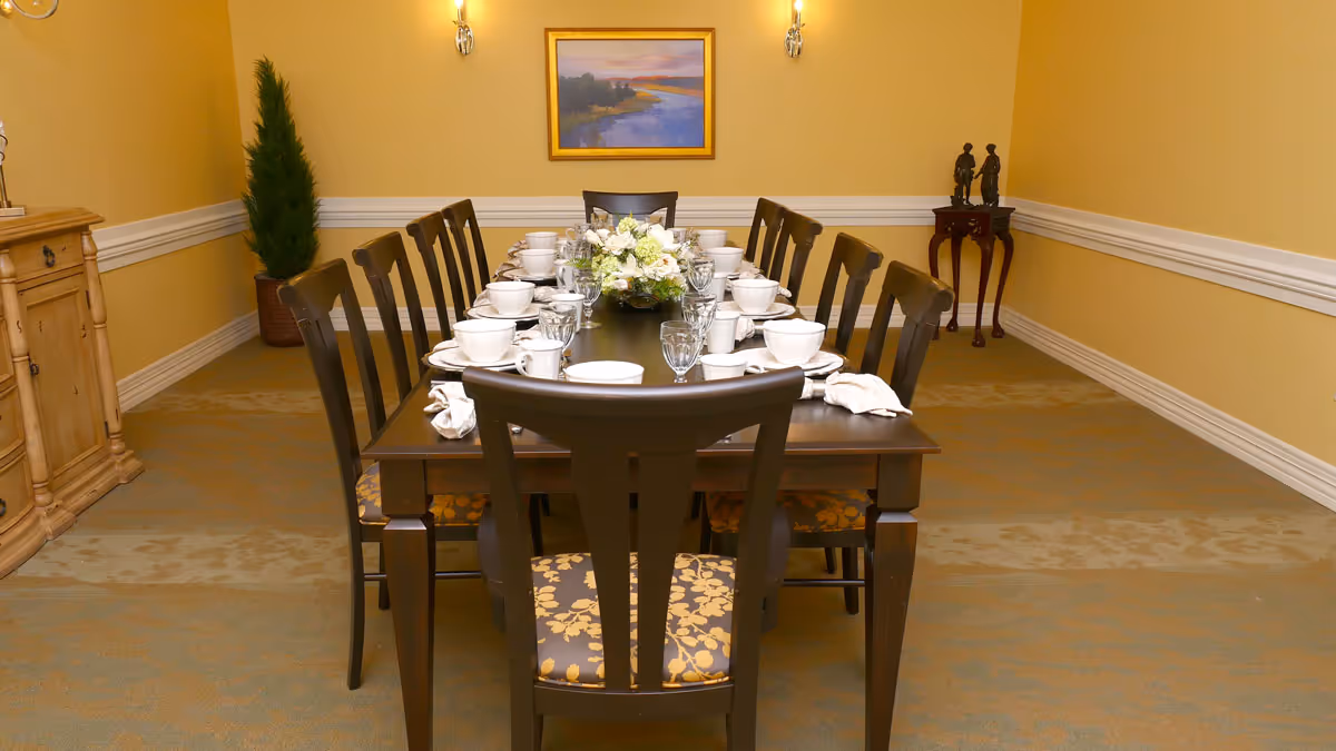 A formal dining room with a long dark wooden table set for a meal with white dishes, cups, and glassware. The table is surrounded by wooden chairs with patterned cushions. The room has yellow walls with white trim, a framed landscape painting on the far wall, a small wooden side table with two statues, a potted plant in the corner, and a wooden cabinet on the left side.