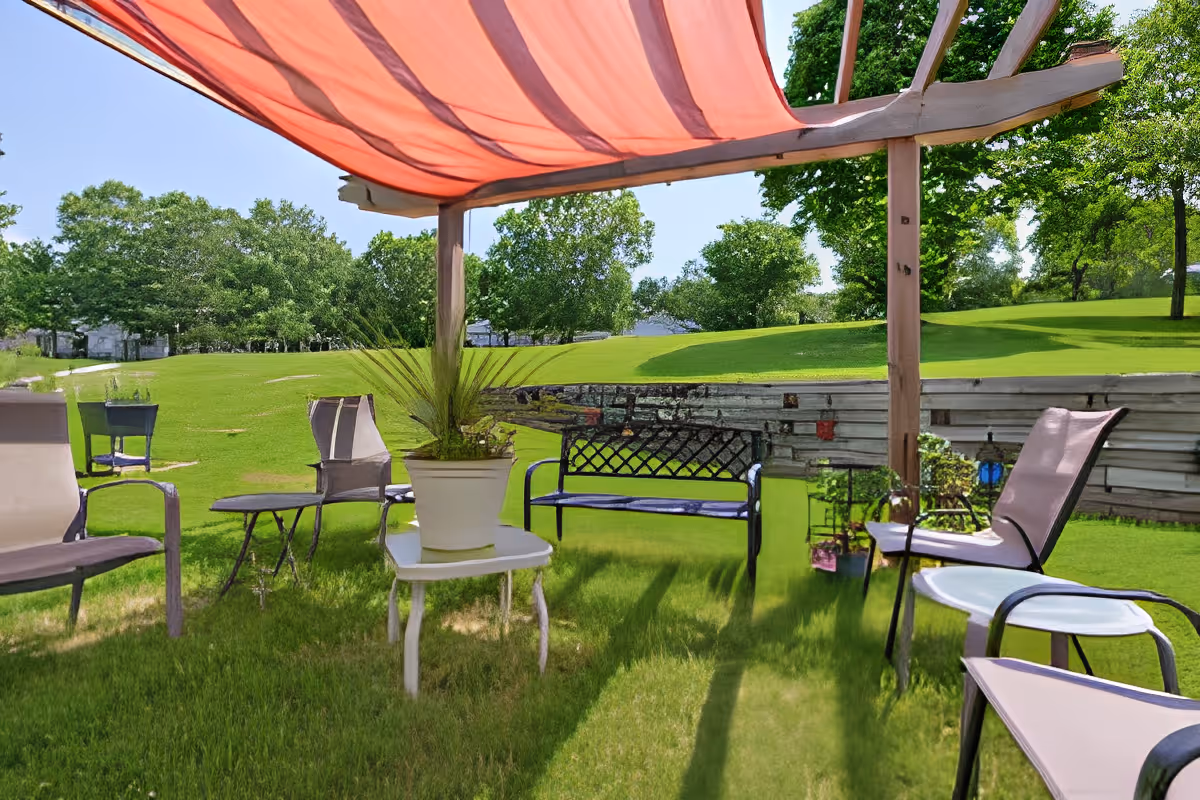 Outdoor shaded seating area with lawn chairs, a bench, tables and a potted plant under a pergola canopy on a grassy lawn.