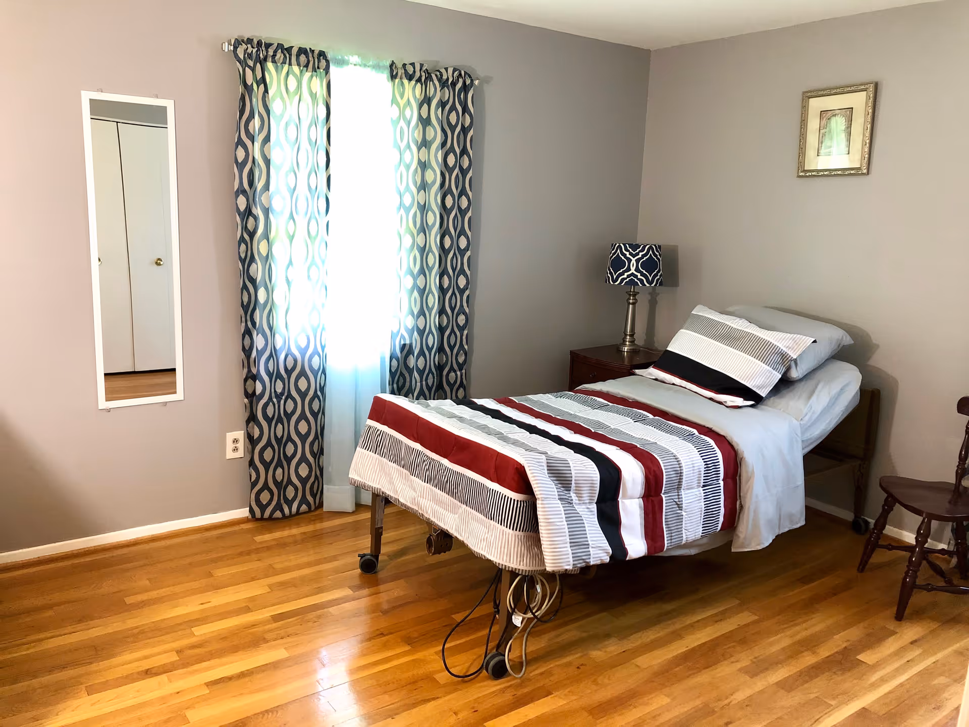 A simple bedroom with a single bed featuring a striped bedspread in red, black, white, and gray. The bed has wheels and is positioned near a nightstand with a blue and white patterned lamp. There is a wooden chair to the right and a window with blue patterned curtains letting in natural light. A small framed picture hangs on the gray wall, and a full-length mirror is mounted on the left wall next to a closet door. The room has hardwood flooring.