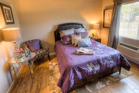 Neatly decorated bedroom with a purple bedspread, tufted headboard, patterned armchair, side tables with lamps, and a window with blinds.