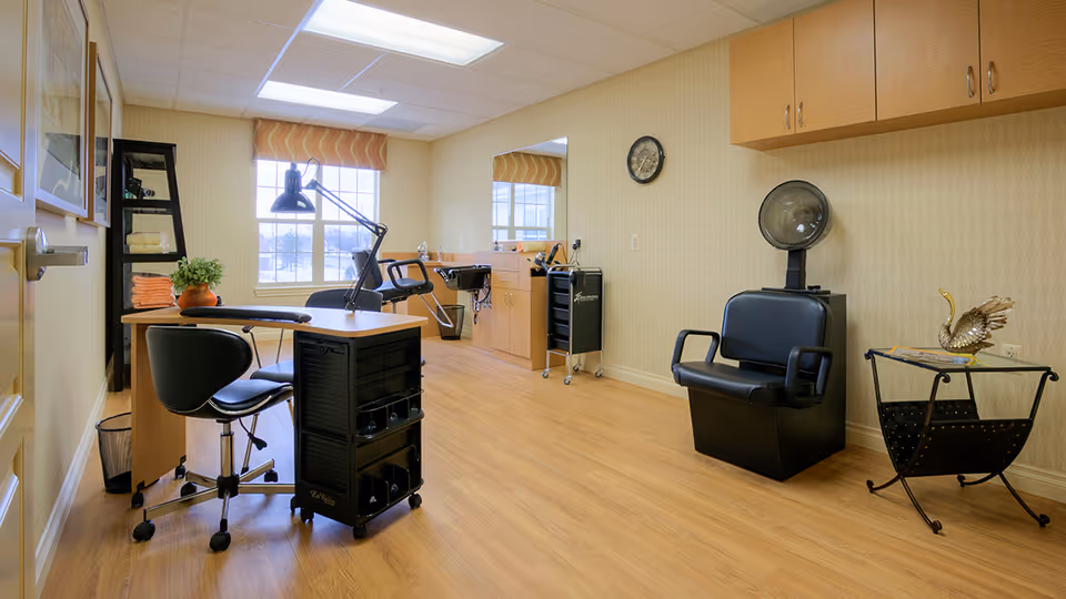 A bright and clean salon room in an assisted living facility featuring a manicure table with two black chairs, a black rolling cart, a hair washing station with a sink and chair, a black hair dryer chair, a small side table with a decorative swan, and a window with a patterned valance letting in natural light.