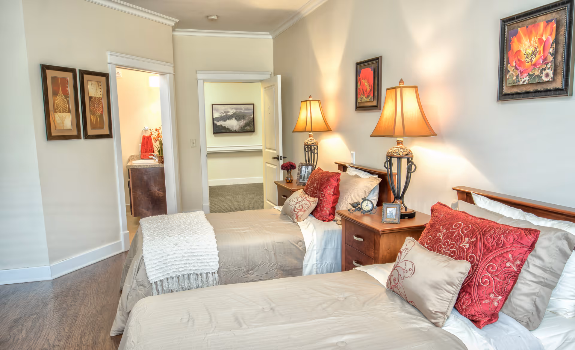 A cozy bedroom with two twin beds featuring beige bedding and red decorative pillows. Each bed has a wooden nightstand with a lamp, a small clock, and framed photos. The walls are adorned with floral artwork, and there is an open door leading to a hallway and a bathroom with a sink and red towels visible.