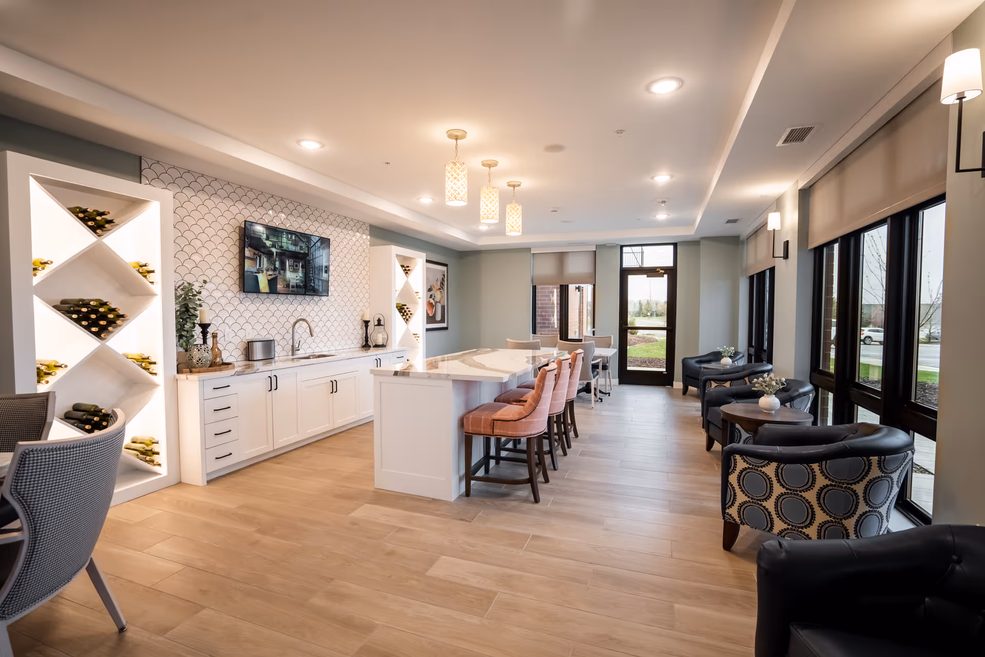 Bright communal dining and lounge area with a central kitchen island, bar stools, wine racks, and large windows.