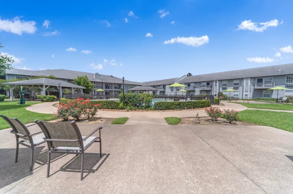 Outdoor courtyard with a fenced swimming pool, patio chairs, umbrellas, a gazebo, flower beds, and a two-story residence under a blue sky.