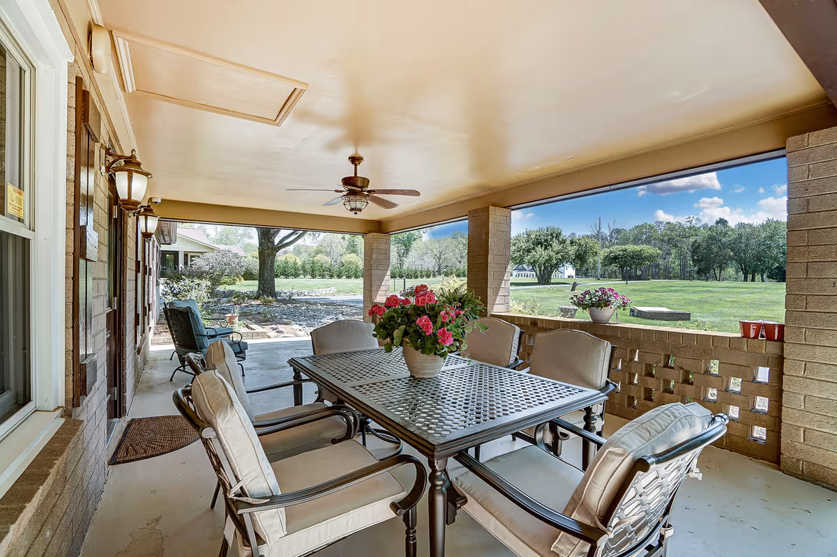 Covered outdoor patio with a metal dining table, cushioned chairs, potted flowers, and a ceiling fan overlooking a grassy yard and trees.