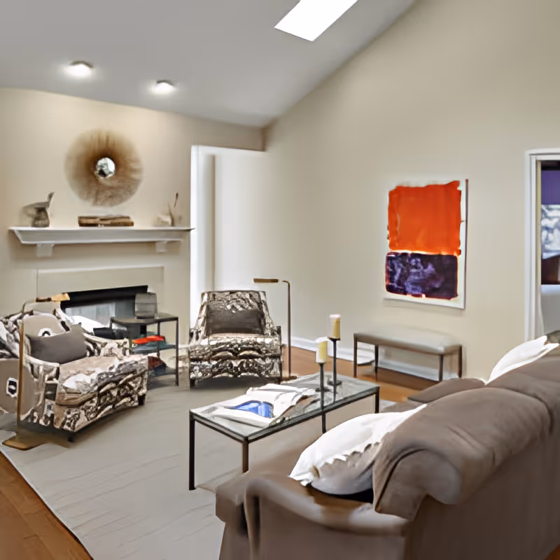 A cozy living room with a brown sofa, two patterned armchairs, a glass coffee table with candles and a book, a fireplace with decorative items on the mantel, and a colorful abstract painting on the wall.