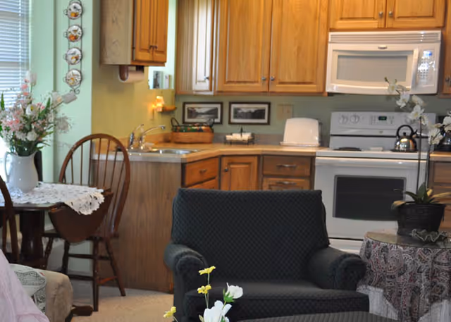 Open-plan interior showing a kitchen with wooden cabinets and white appliances alongside a dining table and upholstered chair.