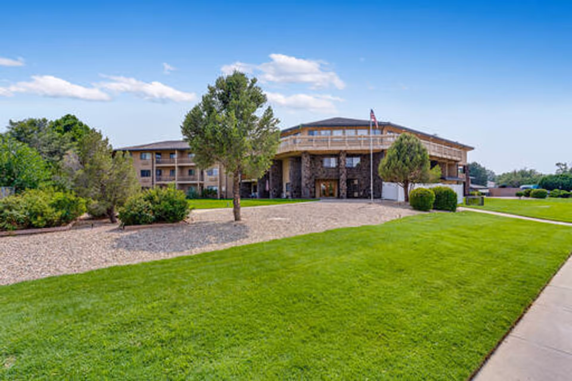 Exterior view of a multi-story senior living facility with a stone entrance, surrounded by green lawns, trees, and bushes under a blue sky with some clouds.