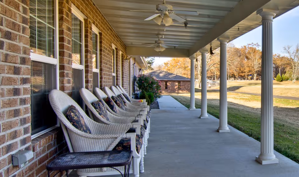 Covered outdoor patio area with a row of white wicker chairs with floral cushions lined up against a brick wall. The patio has white columns supporting the roof and ceiling fans mounted above. In the background, there is a grassy area with trees and another brick building.
