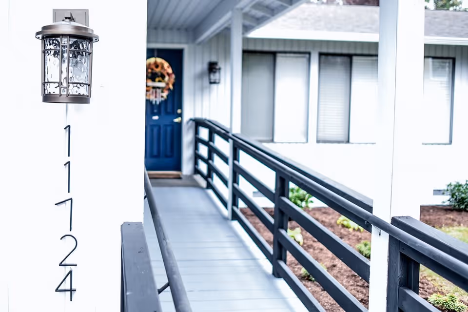 Exterior view of a building entrance with a blue door decorated with a fall wreath. The building has white walls, black railings along a walkway, and the address number 11724 displayed vertically next to a wall-mounted lantern light.