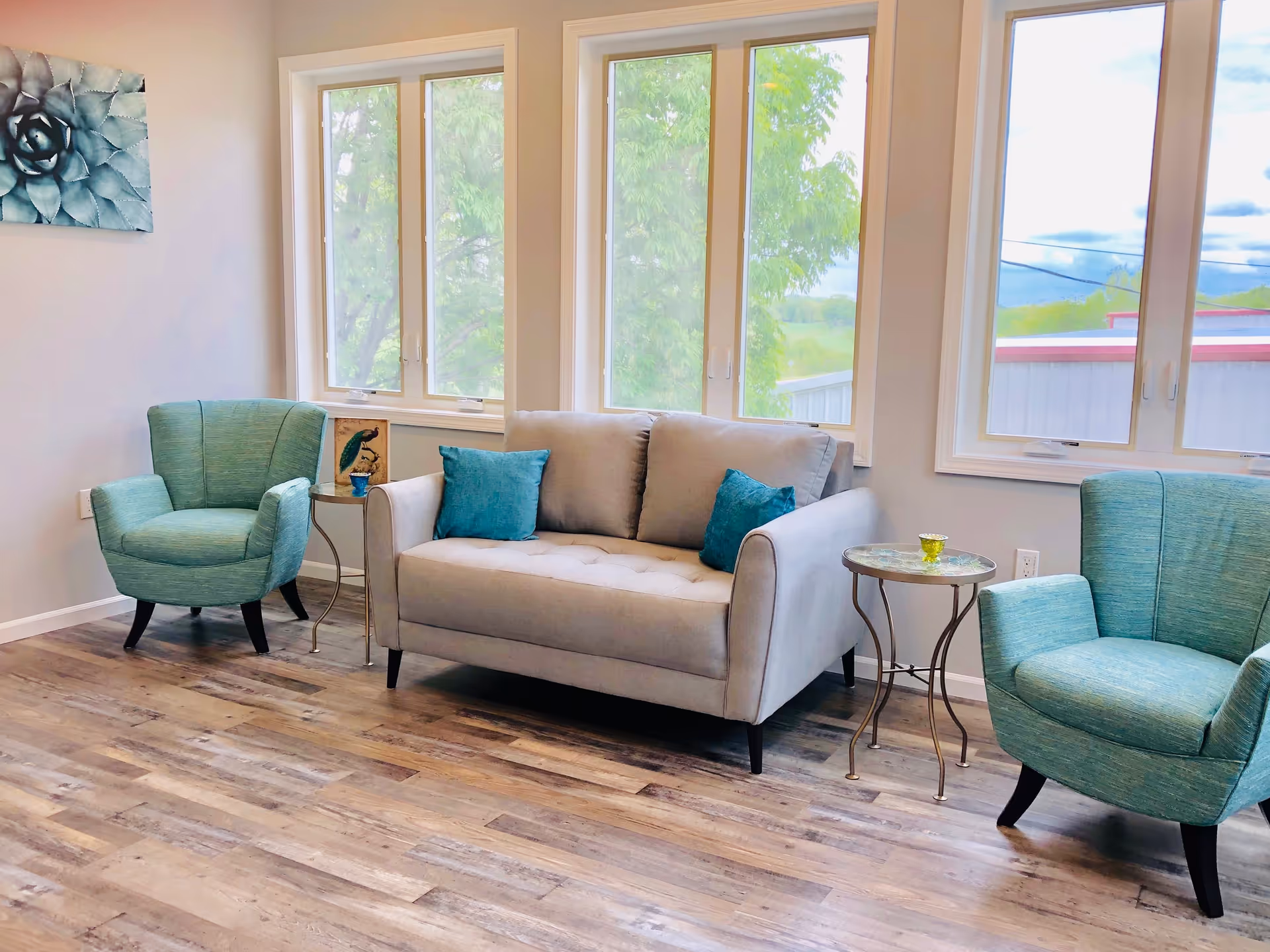 A bright living room with three large windows showing green trees outside. Inside, there is a light gray loveseat with two blue throw pillows, flanked by two teal armchairs. Between the loveseat and each armchair is a small round side table with decorative items on top. The floor is wood with a natural finish, and a blue succulent painting hangs on the wall.