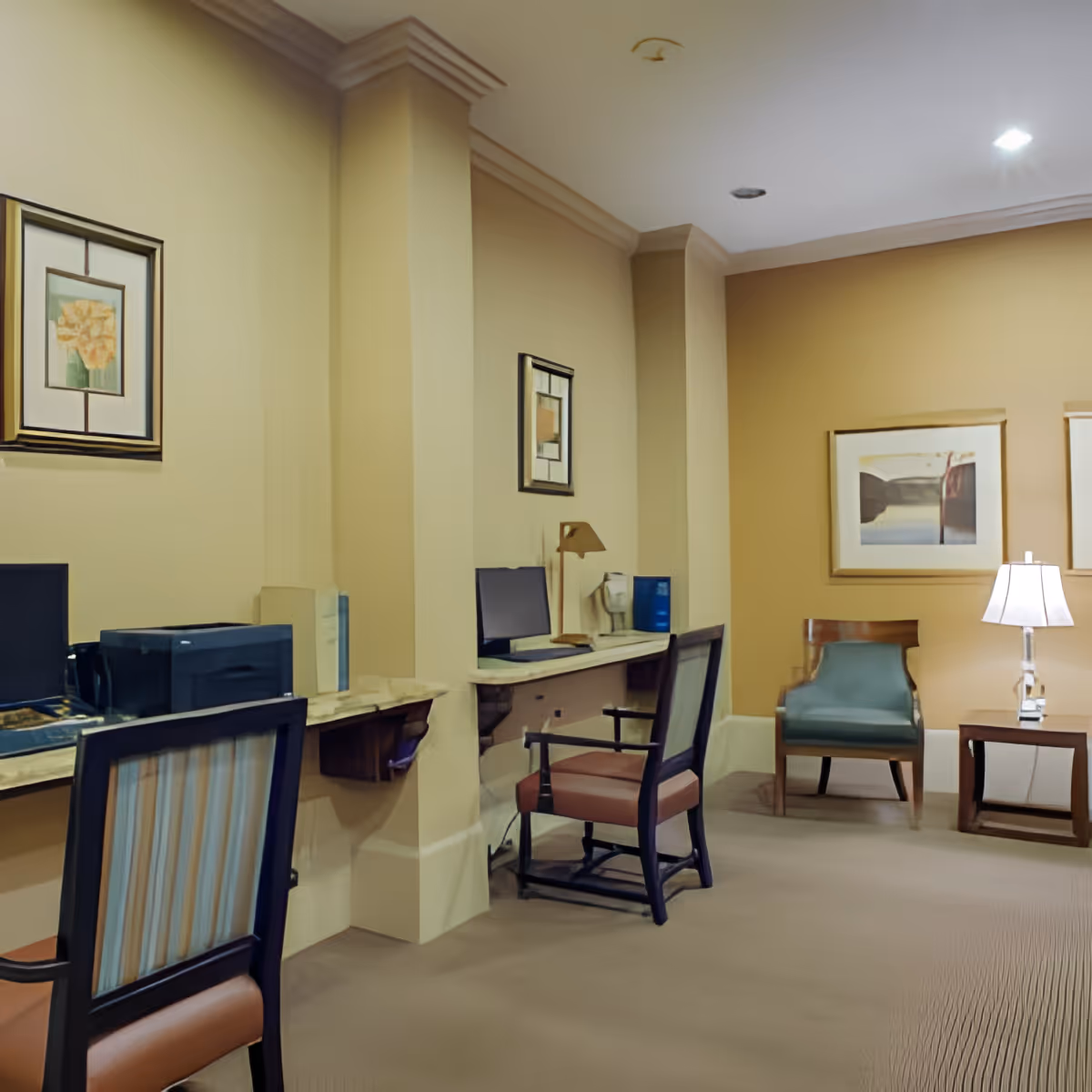 Interior view of a quiet workspace area in a senior living facility with two computer desks, chairs, framed artwork on the walls, and a small seating area with a lamp and armchair.