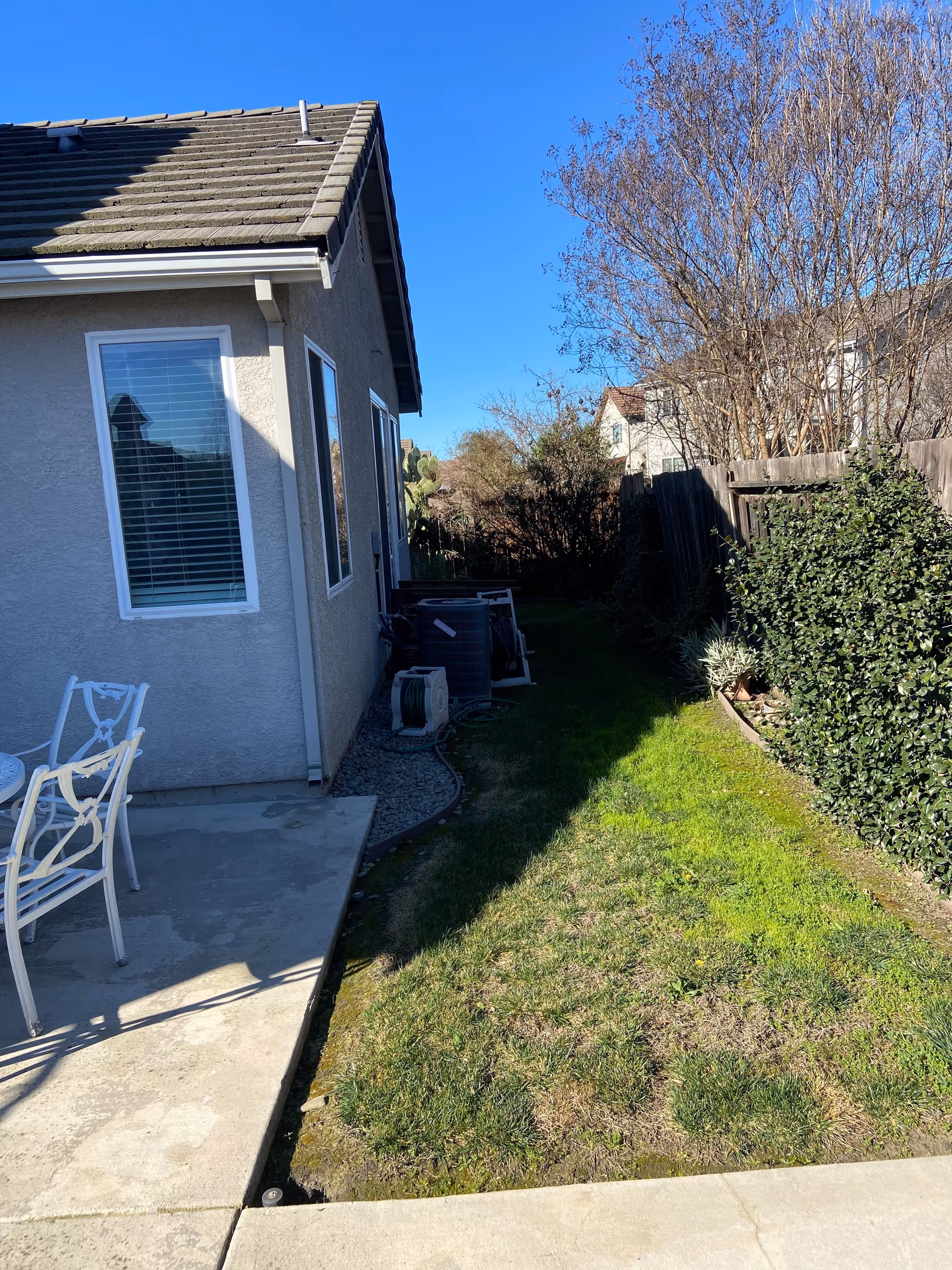 Outdoor side yard area next to a house with a concrete patio, white metal chairs and table, green grass, bushes, and a wooden fence under a clear blue sky.