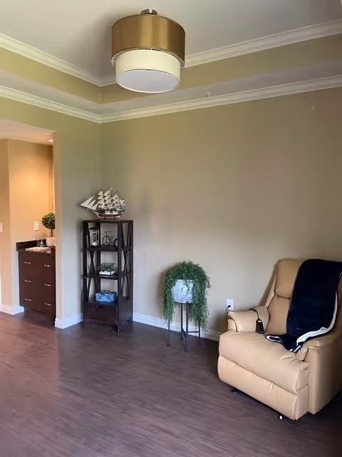 A cozy interior corner of a room with beige walls and dark wood flooring. There is a beige recliner chair with a dark throw blanket on it, a small black metal plant stand holding a green leafy plant, and a dark wooden shelving unit with decorative items including a model ship. A modern ceiling light fixture is visible above, and a doorway leads to a bathroom area with a sink and mirror.