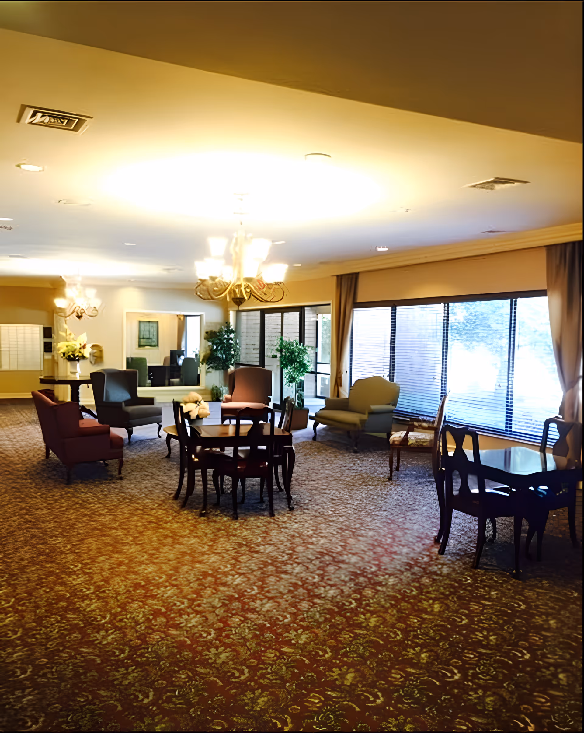 A spacious living room area with patterned carpet, multiple armchairs, and small tables with chairs. Large windows with blinds allow natural light to enter. There are chandeliers hanging from the ceiling and some potted plants near the windows.