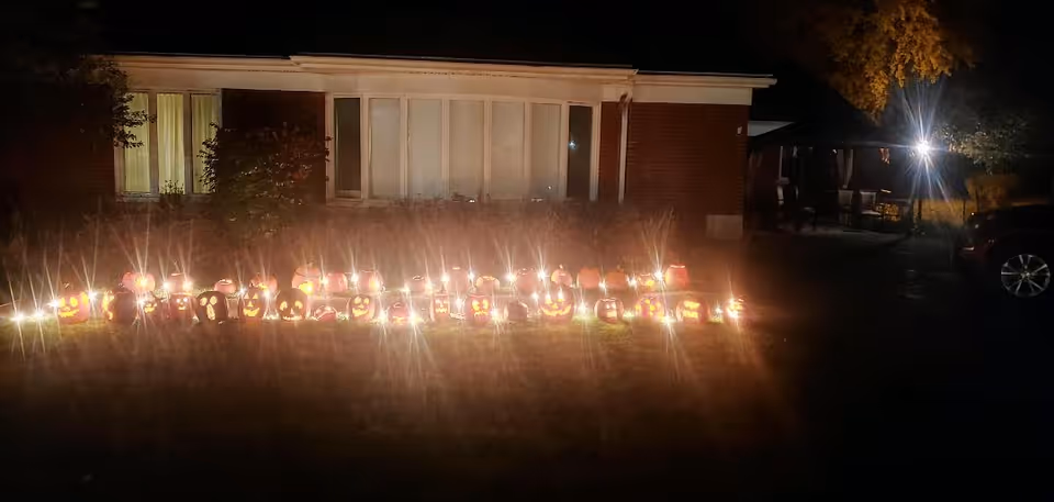 A nighttime outdoor scene showing a row of lit jack-o'-lanterns arranged on the grass in front of a brick building with windows. There is a bright light source illuminating part of the area to the right, where some outdoor furniture and a parked car are visible.