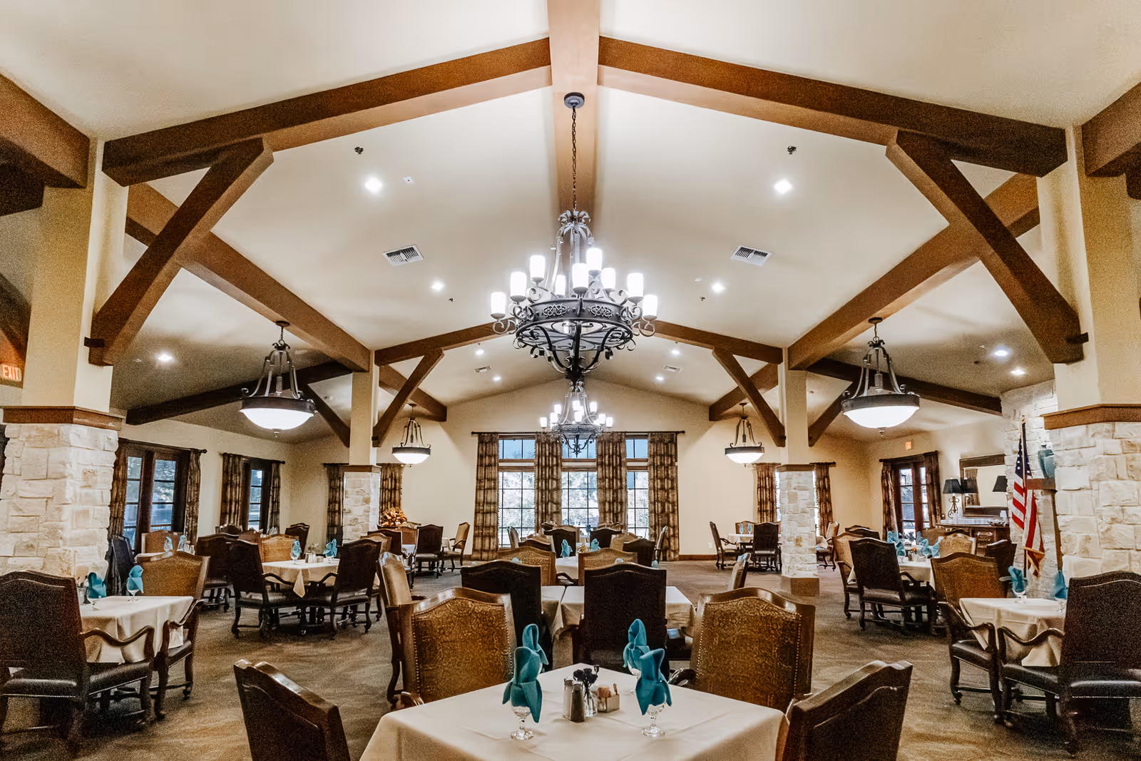Spacious dining room with multiple tables covered in white tablecloths, each set with teal napkins, glasses, and silverware. The room features wooden beams on the ceiling, stone pillars, large windows with patterned curtains, and elegant chandeliers providing warm lighting.