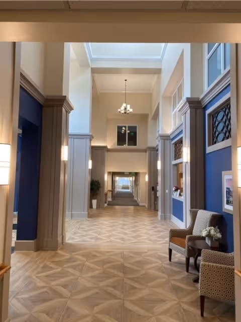 A spacious and well-lit hallway in a senior living facility with high ceilings, beige and blue walls, decorative wall panels, and patterned tile flooring. There are two armchairs with a small table and flowers on the right side, wall sconces providing light, and a chandelier hanging from the ceiling. The hallway extends into the distance with doors and windows visible along the sides.