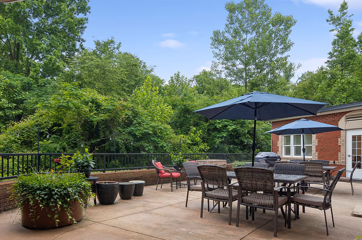 Outdoor patio area with several wicker chairs and tables under large blue umbrellas. There are potted plants along the edge of the patio and lush green trees in the background. A brick building with white-framed windows and a door is visible on the right side.