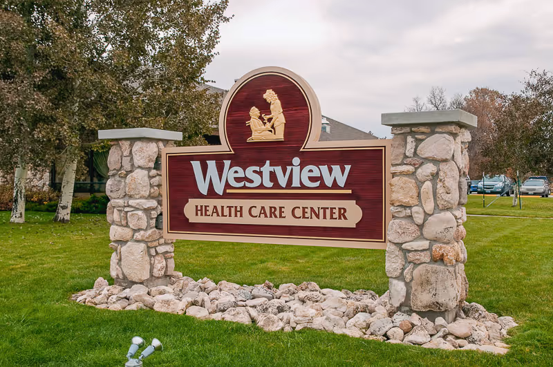 A large stone and wood sign for Westview Health Care Center situated on a grassy area with trees and parked cars in the background under a cloudy sky.