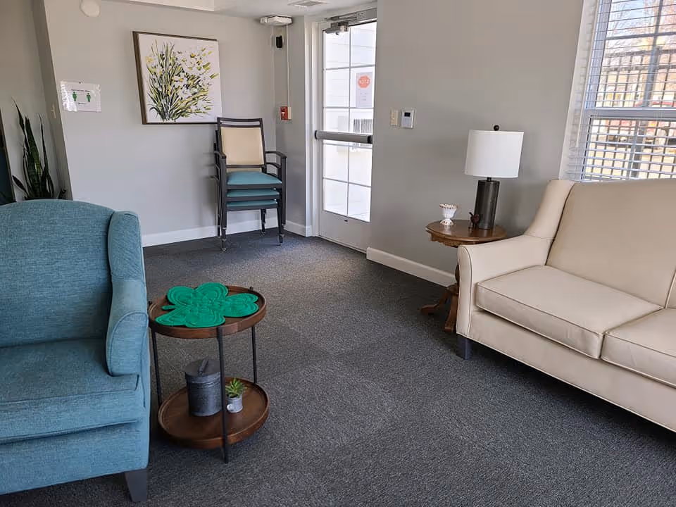A cozy sitting area in a senior living facility with a teal armchair on the left, a small round wooden side table with a green decorative item and a small plant, and a beige loveseat on the right next to a wooden side table with a lamp. In the background, there is a glass door, a stack of chairs, a wall painting of flowers, and a window with blinds letting in natural light.