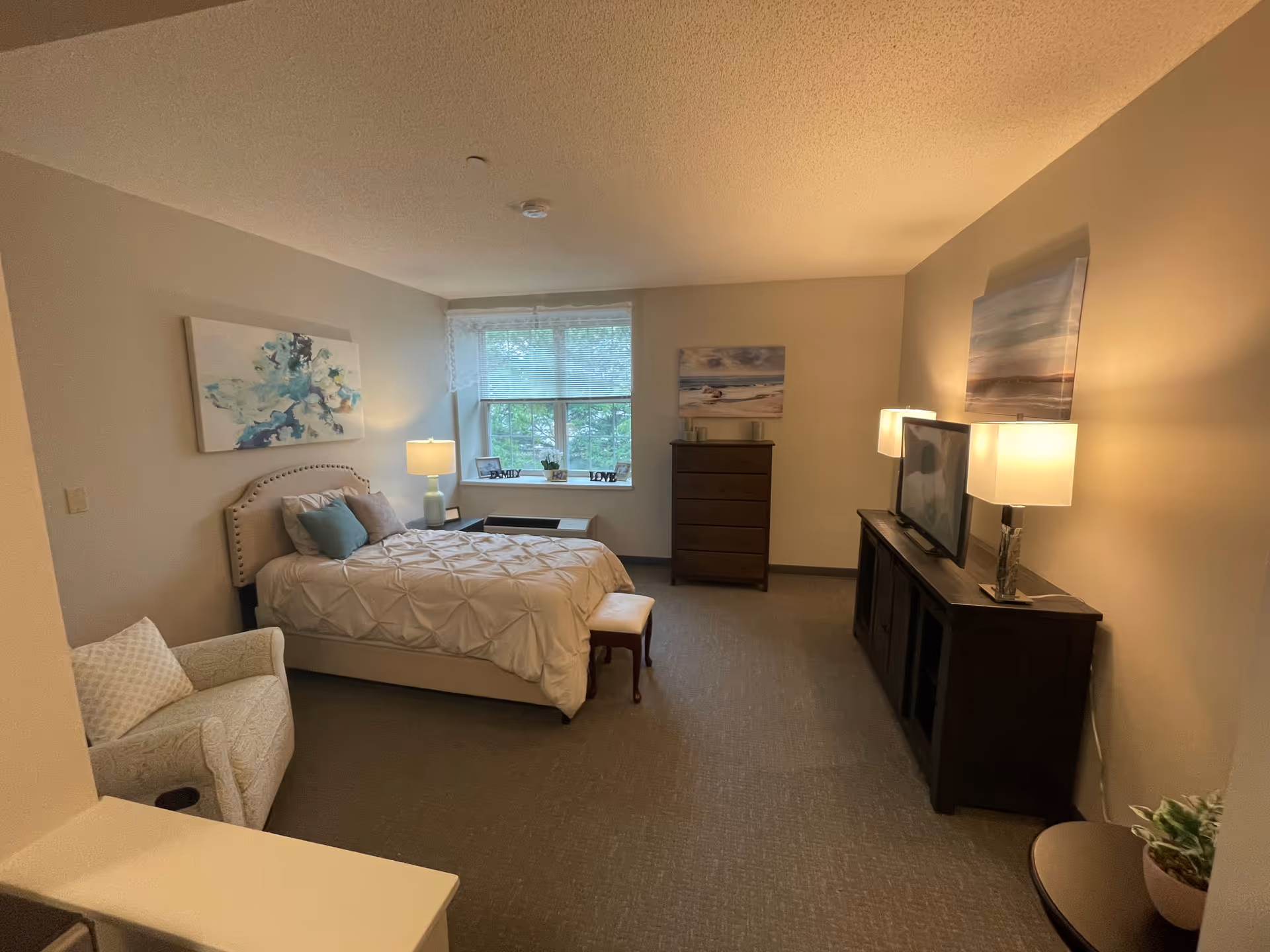 Furnished senior suite bedroom with a bed, armchair, dresser, TV console, and lamps near a window.
