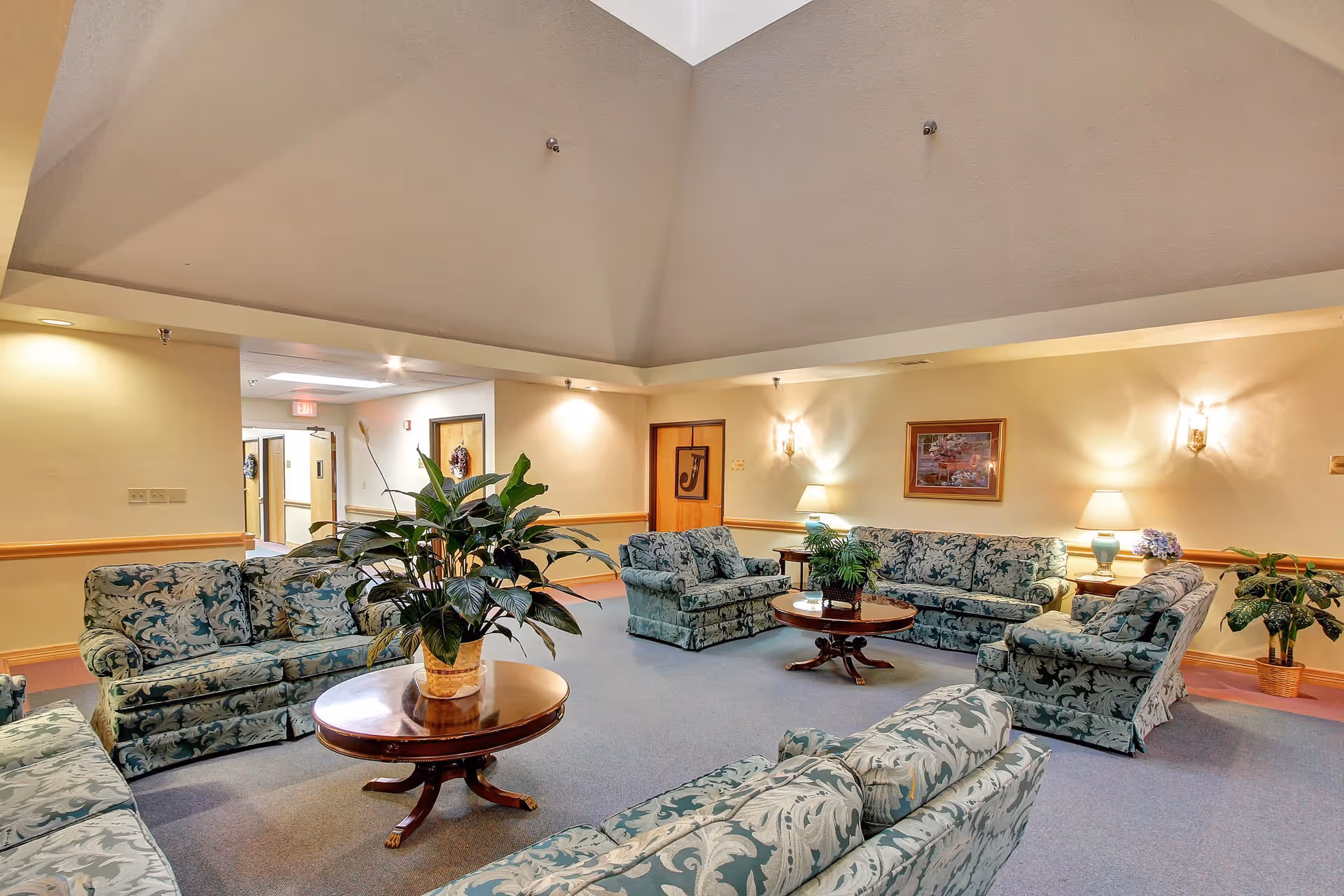A spacious living room area in a senior living facility with multiple patterned sofas arranged around two wooden coffee tables, each with a potted plant. The room has beige walls with wall sconces and table lamps providing warm lighting, a framed picture on the wall, and a high ceiling. There are doors and a hallway visible in the background.