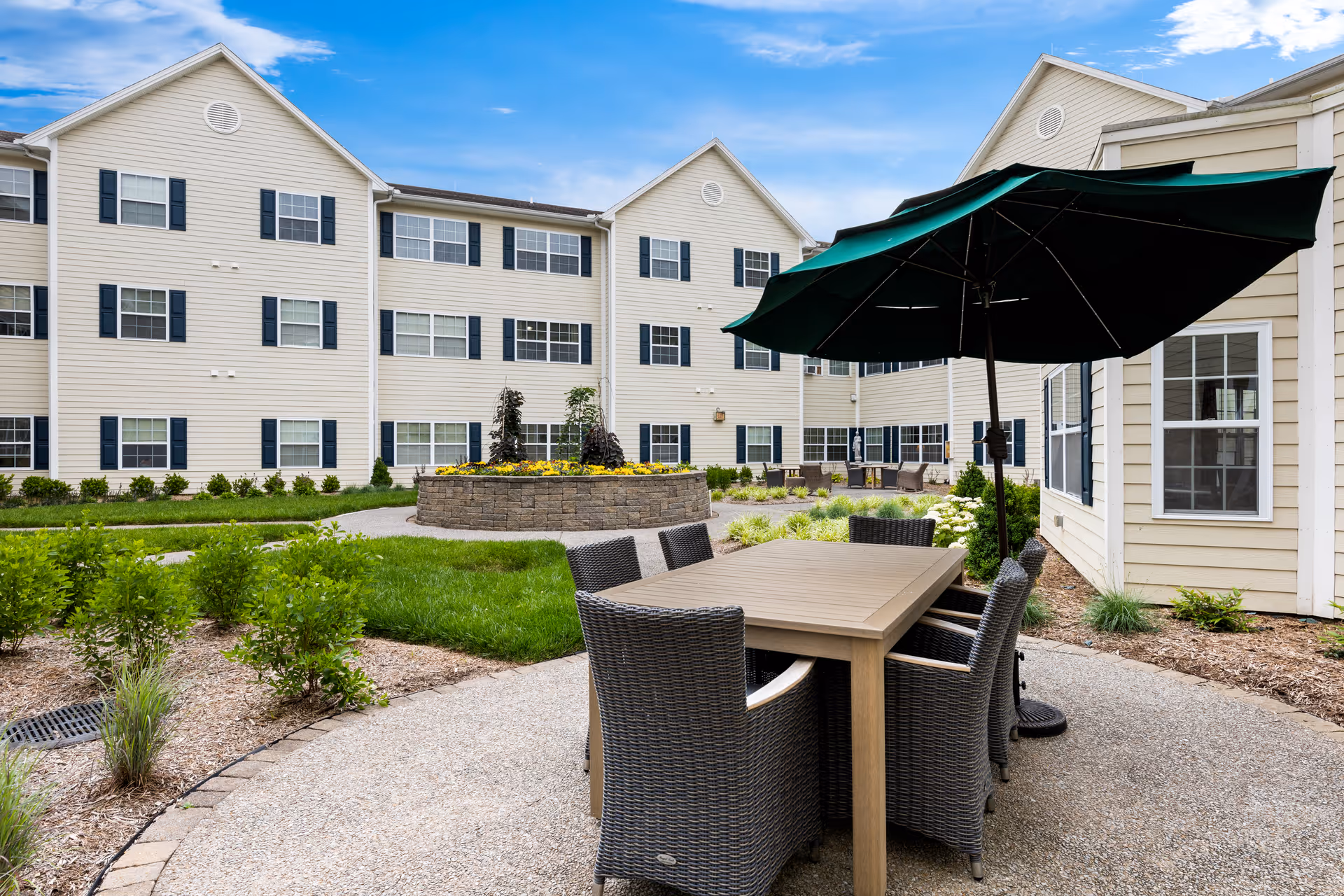 Outdoor courtyard area at Vitality Living Hendersonville featuring a patio table with six wicker chairs and a large green umbrella. The courtyard is surrounded by a three-story beige building with multiple windows and black shutters. There is a circular raised flower bed with yellow flowers and greenery in the center of the courtyard, along with well-maintained landscaping and a paved walkway.