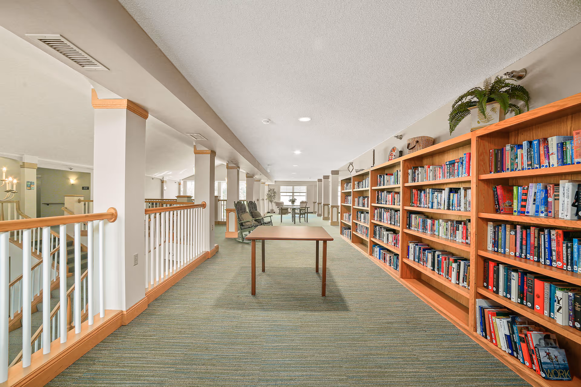 A bright and spacious indoor library area with wooden bookshelves filled with books on the right side, a table in the center, and several chairs near the windows at the far end. The space has light-colored walls, carpeted flooring, and white railings along the left side overlooking a lower level.