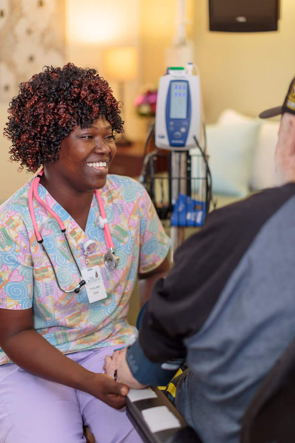 A smiling female nurse wearing colorful scrubs and a pink stethoscope is holding hands and talking with an elderly man seated in a wheelchair in a warmly lit room with medical equipment in the background.