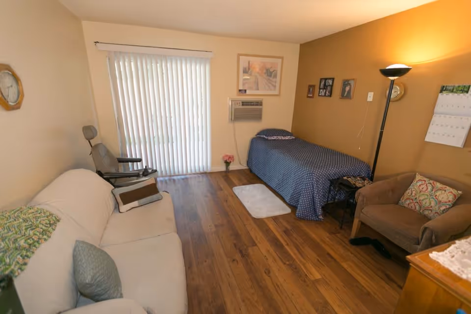 A cozy senior living room with a single bed covered in a blue polka dot blanket, a beige couch with pillows, a cushioned armchair, a floor lamp, and a sliding glass door with vertical blinds. The room has wooden flooring, light brown walls, framed pictures, a wall-mounted air conditioner, and a calendar on the wall.
